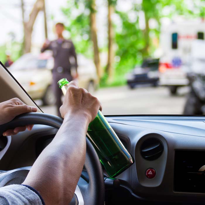 Man holding alcohol bottle at the wheel with cop in background and wrecked vehicles